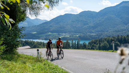 Zwei Radfahrer auf einer Landstraße mit Berg- und Seehintergrund