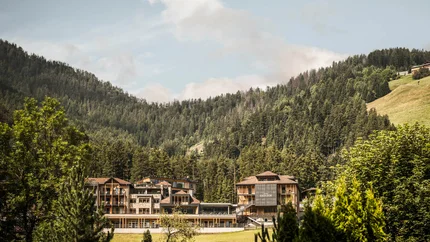 Excelsior Dolomites Life Resort © Hannes Niederkofler Hotelgebäude eingebettet in grüne Berge und Wald unter blauem Himmel