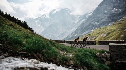 Zwei Radfahrer fahren auf Bergstraße mit schneebedeckten Bergen im Hintergrund