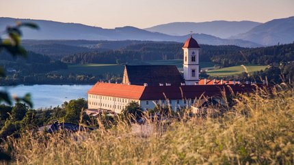 Klostergebäude mit rotem Dach vor Berg- und Seenlandschaft bei Sonnenuntergang