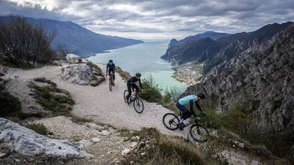 Nördlicher Gardasee © Gijs Ferkranus - Wielrenblad Magazine Drei Radfahrer auf einem Bergweg mit Blick auf einen See und Berge